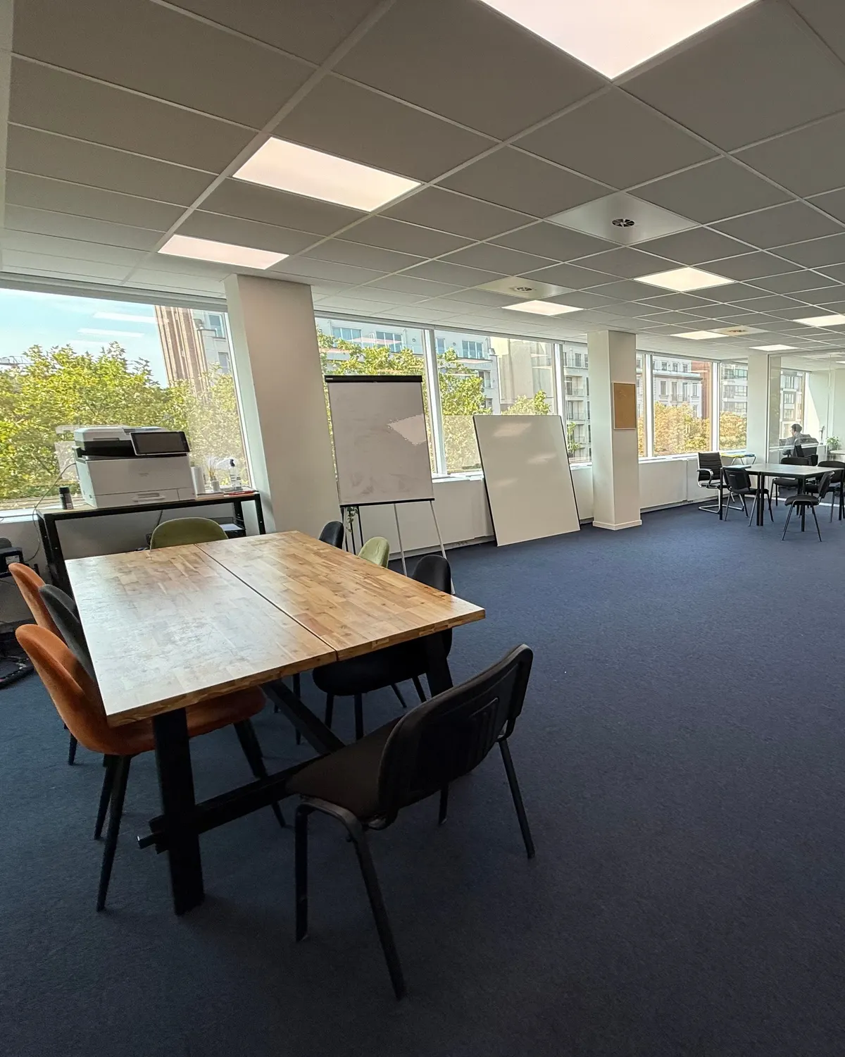 Grote, heldere kantoorverdieping met een houten tafel, stoelen, blauw tapijt en achteraan een vergaderzaal met glazen wanden